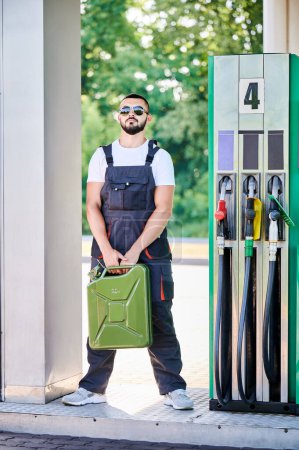 Photo for Professional worker holding canister in hands. Confident operator in sunglasses and overalls waiting for customers. Male worker standing on background of gas station with cistern in hands. - Royalty Free Image