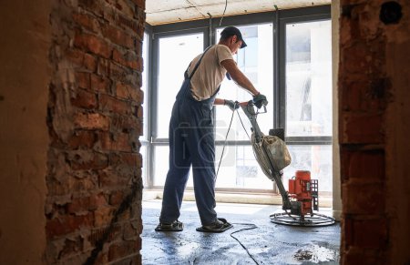 Full length of male worker using troweling machine while screeding floor in apartment under renovation. Man finishing concrete surface with floor screed grinder machine in room with large window.