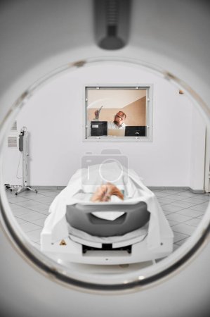 Foto de Tomografía computarizada médica o escáner de resonancia magnética. Médico examinando los resultados de la resonancia magnética, paciente mujer acostada en la litera. Médico con gafas, con resultados de resonancia magnética, mirando, estudiando, pensando. - Imagen libre de derechos
