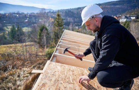Foto de Carpenter hammering nail into OSB panel on the roof top of future cottage. Man worker building wooden frame house. Carpentry and construction concept. - Imagen libre de derechos