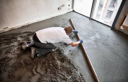 Männliche Bauarbeiter legen Estrichschienen auf den Boden, der mit Sand-Zement-Gemisch bedeckt ist. Mann glättet und nivelliert Fläche mit gerader Kante beim Estrichboden in Wohnung.