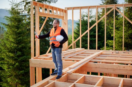 Photo for Manager overseeing construction of wooden two-story house located close to woods. Bearded man in glasses studying construction plan. Objective is to implement modern ecological construction methods. - Royalty Free Image