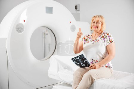 Foto de Tomografía computarizada médica o escáner de resonancia magnética. Retrato de paciente femenina haciendo resonancia magnética. Mujer rubia sosteniendo el resultado de la resonancia magnética, escanear, sonriendo a la cámara, mostrando los pulgares hacia arriba. Concepto de medicina y asistencia sanitaria. - Imagen libre de derechos