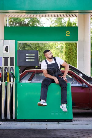 Photo for Young gas station operator waiting for new customers. Professional worker sitting next to gasoline column ready to work. Male adult in overalls sitting at gas station and looking away. - Royalty Free Image