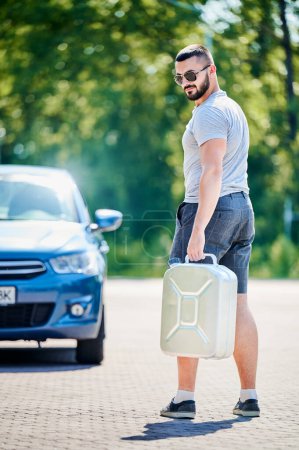 Photo for Young male looking for petrol station to fill canister. Man trying to find nearest gas station. Adult man in casual clothes holding canister on background of blue car. - Royalty Free Image