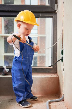 Téléchargez les photos : Kid welding electrical wire with electric soldering iron. Child electrician in work overalls and safety helmet installing electrical cables and sockets in apartment under renovation. - en image libre de droit