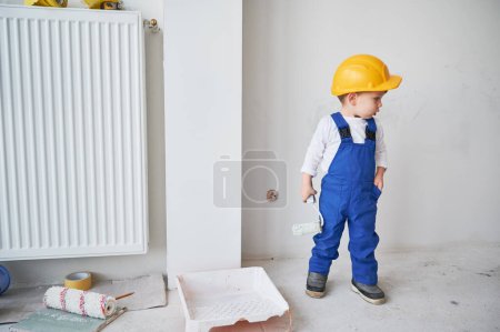 Volle Länge der Bauarbeiter Malerei Wand in Wohnung in Renovierung. Kind im Schutzhelm und Arbeitsanzug mit Farbroller beim Spielen zu Hause.