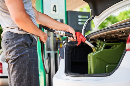Photo for Male driver with gas pump refueling canister with fuel in car trunk. Man holding hose and filling cistern with gasoline. Close view of man filling canister on background of gasoline column. - Royalty Free Image