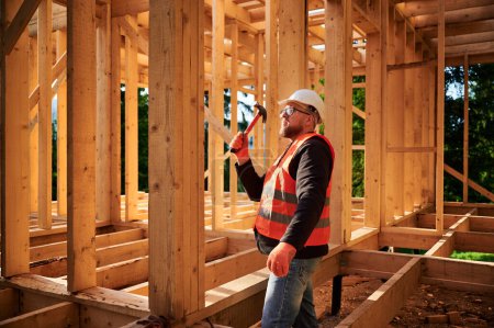 Photo for Carpenter constructing wooden frame house. Bearded man with glasses is hammering nails into the structure, wearing protective helmet and construction vest. Concept of modern ecological construction. - Royalty Free Image