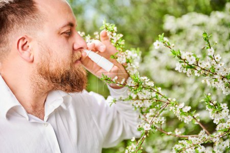 Téléchargez les photos : Homme allergique à l'aide de gouttes nasales médicales, souffrant d'allergie saisonnière au printemps dans le jardin en fleurs. Un bel homme soignant son nez qui coule devant un arbre en fleurs à l'extérieur. Concept d'allergie printanière. - en image libre de droit