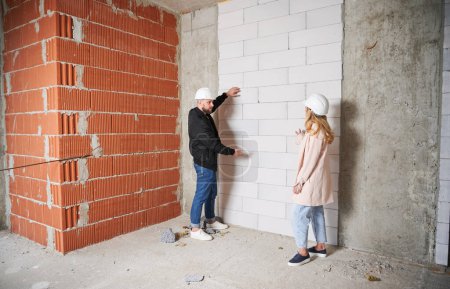 Photo for Full length of woman standing by brick wall and discussing apartment replanning and renovation with specialist. Female homeowner having meeting with builder in building under construction. - Royalty Free Image
