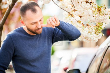 Männerallergiker leiden unter saisonaler Allergie im Frühling im blühenden Garten im Frühling. Ein bärtiger junger Mann niest und pustet laufende Nase vor einem blühenden Baum. Frühjahrsallergiekonzept.