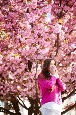 Woman allergic suffering from seasonal allergy at spring in blossoming garden at springtime. Woman sneezing and blowing nose using nasal handkerchief in front of blooming tree. Spring allergy concept.