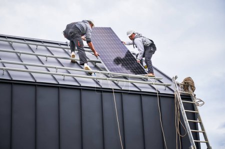 Foto de Constructores instalando sistema de panel solar en el techo de la casa. Hombres trabajadores en cascos que llevan módulo solar fotovoltaico al aire libre. Concepto de energía alternativa y renovable. - Imagen libre de derechos