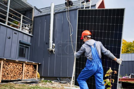 Foto de Montaje instalación de sistema de panel solar en el techo de la casa. Hombre trabajador en casco llevando módulo solar fotovoltaico al aire libre. Concepto de energía alternativa y renovable. - Imagen libre de derechos