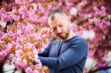 Téléchargez les photos : Homme allergique souffrant d'allergie saisonnière au printemps dans le jardin en fleurs au printemps. Homme barbu sentant des démangeaisons des yeux devant un arbre en fleurs. Concept d'allergie printanière. - en image libre de droit