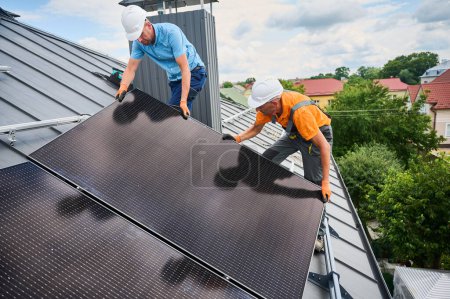Foto de Los trabajadores que construyen el sistema de paneles solares en la azotea de la casa. Dos hombres instaladores en cascos instalando módulo solar fotovoltaico al aire libre. Concepto de generación de energía alternativa, verde y renovable. - Imagen libre de derechos