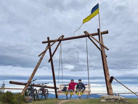 Two men resting from biking, sitting on large wooden swing outdoor near two electric mountain bikes. Carpathians, Ukraine.