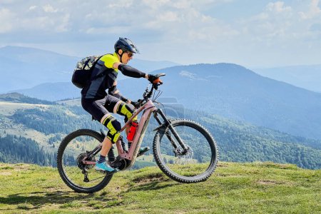 Cyclist man riding electric mountain bike outdoors. Male tourist biking along grassy trail in the mountains, wearing helmet and backpack. Concept of sport, active leisure and nature.