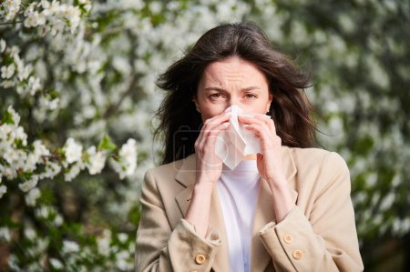 Photo for Woman allergic suffering from seasonal allergy at spring in blossoming garden at springtime. Young woman sneezing and blowing nose with nasal handkerchief in front of blooming tree. Allergy concept. - Royalty Free Image