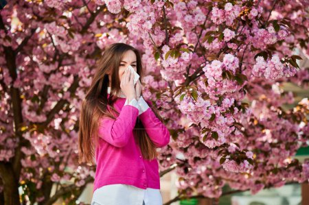 Photo for Woman allergic suffering from seasonal allergy at spring in blossoming garden at springtime. Woman sneezing and blowing nose using nasal handkerchief in front of blooming tree. Spring allergy concept. - Royalty Free Image