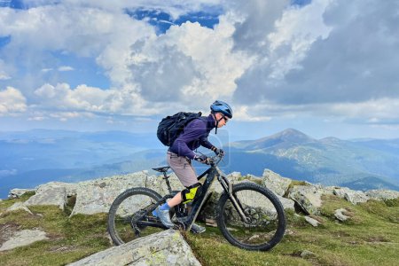 Hombre ciclista montando bicicleta de montaña eléctrica al aire libre. Ciclismo turístico masculino a lo largo de sendero herboso en las montañas, con casco y mochila. Concepto de deporte, ocio activo y naturaleza.