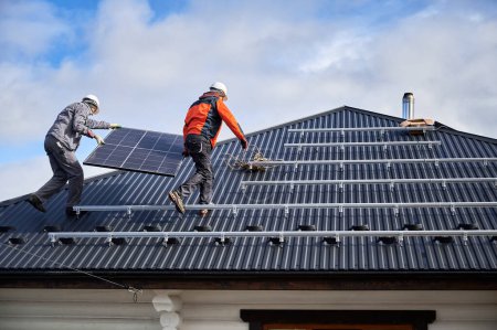 Foto de Los trabajadores que llevan panel solar en un techo de la casa. Dos ingenieros instalando la batería solar en el día soleado. Recursos energéticos alternativos. - Imagen libre de derechos