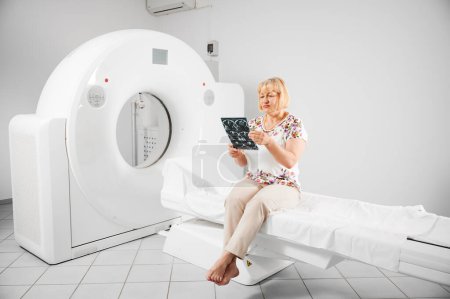 Medical computed tomography or MRI scanner. Patient making MRI, sitting on couchette. Blonde woman holding MRI result, scan, x-ray, looking, examining. Concept of medicine and healthcare.