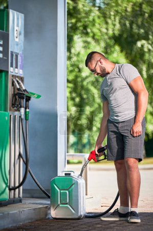 Photo for Handsome driver refueling canister with gasoline. Man filling cistern with fuel in case of unforeseen circumstances. Man in casual clothes with pump nozzle on background of modern gas station. - Royalty Free Image