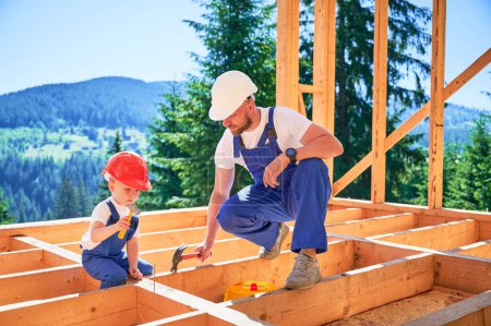 Photo for Father with toddler son building wooden frame house. Male builder teaching his son the technique of pounding nails with hammer on construction site, wearing helmets and blue overalls on sunny day. - Royalty Free Image