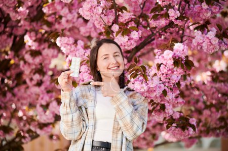 Frauen, die allergisch sind, leiden unter saisonaler Allergie im Frühling. Junge glückliche Frau zeigt Packung Pillen, posiert im blühenden Garten im Frühling. Antihistaminisches Medikamentenkonzept