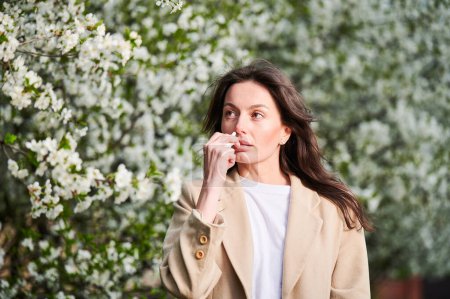 Photo for Woman allergic using medical nasal drops, suffering from seasonal allergy at spring in blossoming garden. Young woman treating runny nose in front of blooming tree outdoors. Spring allergy concept. - Royalty Free Image