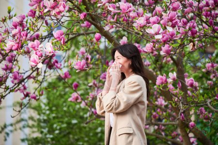 Photo for Woman allergic suffering from seasonal allergy at spring in blossoming garden at springtime. Young woman sneezing and blowing nose with nasal handkerchief in front of blooming tree. Allergy concept. - Royalty Free Image