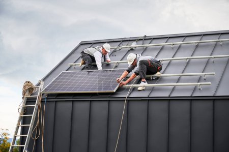 Foto de Montajes instalando paneles solares fotovoltaicos en el techo de la casa. Los hombres ingenieros en cascos que construyen el sistema del módulo solar con la ayuda de la llave hexagonal. Concepto de energía alternativa y renovable. - Imagen libre de derechos