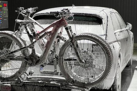 Vehículo eléctrico y bicicletas en auto-servicio de lavado de coches durante la noche, cubierto de espuma, lo que indica que está en el medio de un ciclo de lavado. Bicicletas montadas en un portaequipajes en la parte trasera.