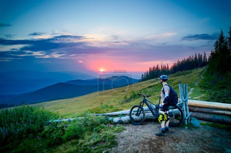 Cyclist man riding electric bike outdoors in the evening. Male tourist resting on grassy hill, enjoying beautiful mountain landscape, wearing helmet and backpack. Concept of active leisure.