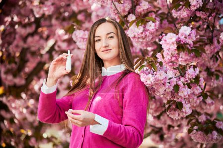 Photo for Woman allergic using medical nasal drops, suffering from seasonal allergy in blossoming garden. Portrait of smiling woman holding nasal spray near blooming tree. Spring allergy concept. - Royalty Free Image