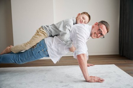Energetic father and his little son training at home. Kid climbed dads back while he doing plank. Young family enjoying activity games at home.