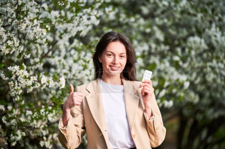 Photo for Woman allergic suffering from seasonal allergy at spring. Young happy woman showing allergy drugs, giving thumbs up, posing in blossoming garden at springtime. Antihistamine medication concept - Royalty Free Image