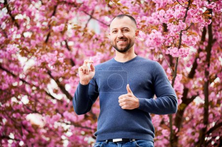 Photo for Man allergic using medical nasal drops, suffering from seasonal allergy at spring in blossoming garden. Portrait of smiling man showing thumbs up near blooming tree outdoors. Spring allergy concept. - Royalty Free Image
