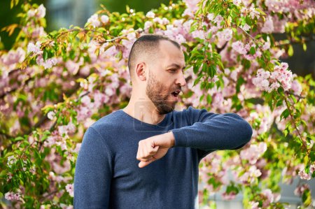 Männerallergiker leiden unter saisonaler Allergie im Frühling im blühenden Garten im Frühling. Ein bärtiger junger Mann niest und hat eine laufende Nase vor einem blühenden Baum. Frühjahrsallergiekonzept.