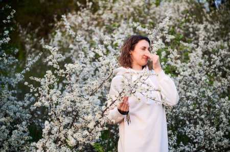 Woman allergic suffering from seasonal allergy at spring in blossoming garden at springtime. Young woman sneezing, closing nose by hand in front of blooming tree. Spring allergy concept