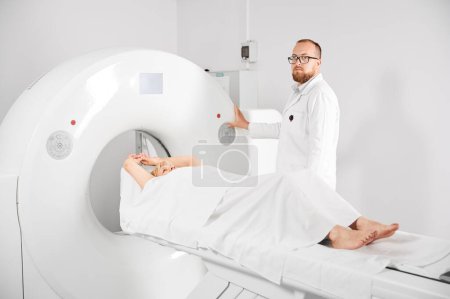 Foto de Tomografía computarizada médica o escáner de resonancia magnética. Radiólogo masculino presiona botón de resonancia magnética para examinar paciente femenino, mirando a la cámara. - Imagen libre de derechos