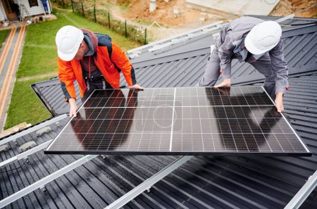 Foto de Electricistas instalando sistema de panel solar en el techo de la casa. Hombres trabajadores en cascos que llevan módulo solar fotovoltaico al aire libre. Concepto de energía alternativa y renovable. - Imagen libre de derechos