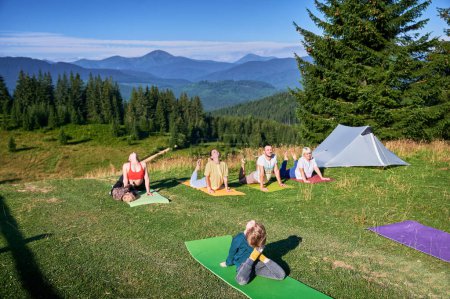 Eine Gruppe von Yoga-Teilnehmern posiert im Freien beim Zelten in den Bergen. Erwachsene und Kinder auf Yogamatten, die jeweils morgens unter strahlend blauem Himmel eine Yoga-Pose hinlegen. Junge ist Ausbilder.
