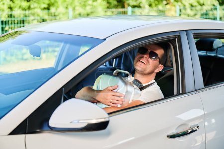 Glücklicher Mann umarmt Kanister voller Benzin. Der Mensch ist froh, dass er Kanister genommen hat und zusätzlichen Treibstoff für unvorhergesehene Umstände hat. Lächelnder Mann mit Sonnenbrille sitzt in weißem Auto und hält Zisterne.