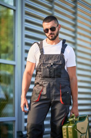 Strong gas station worker holding canister full of fuel. Portrait of male operator caring heavy cistern with gasoline. Confident man in sunglasses and overalls with canister.
