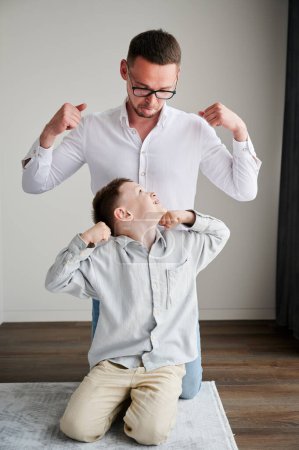 Joven padre jugando con su curioso hijo. Chico gracioso mostrando músculos a su padre. Familia disfrutando de juegos de actividad en casa.