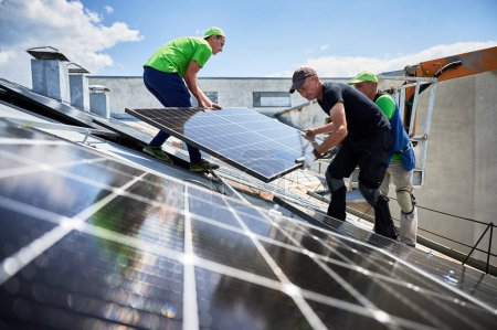 Los trabajadores que construyen el sistema de paneles solares en la azotea de metal de la casa con la ayuda de grúa elevadora. Dos instaladores de hombres que llevan módulo solar fotovoltaico al aire libre. Concepto de generación de energía renovable.
