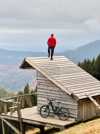 Person resting on edge of steeply sloped wooden rooftop in mountainous area after riding on electric mountain bike on cloudy day. Below the rooftop, there is wooden deck with mountain bicycle.